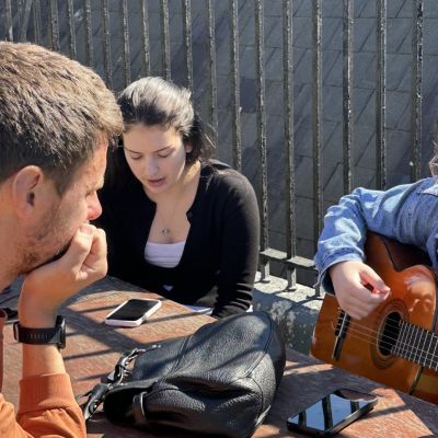 A tutor listens to two Music Makers students practicing a song they've been working on for the workshop. The three sit on a sunny rooftop terrace.